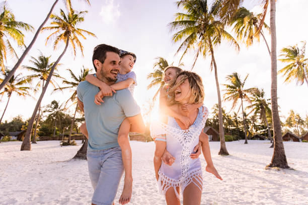 Happy parents and their small kids having fun while piggybacking on the beach in summer day.